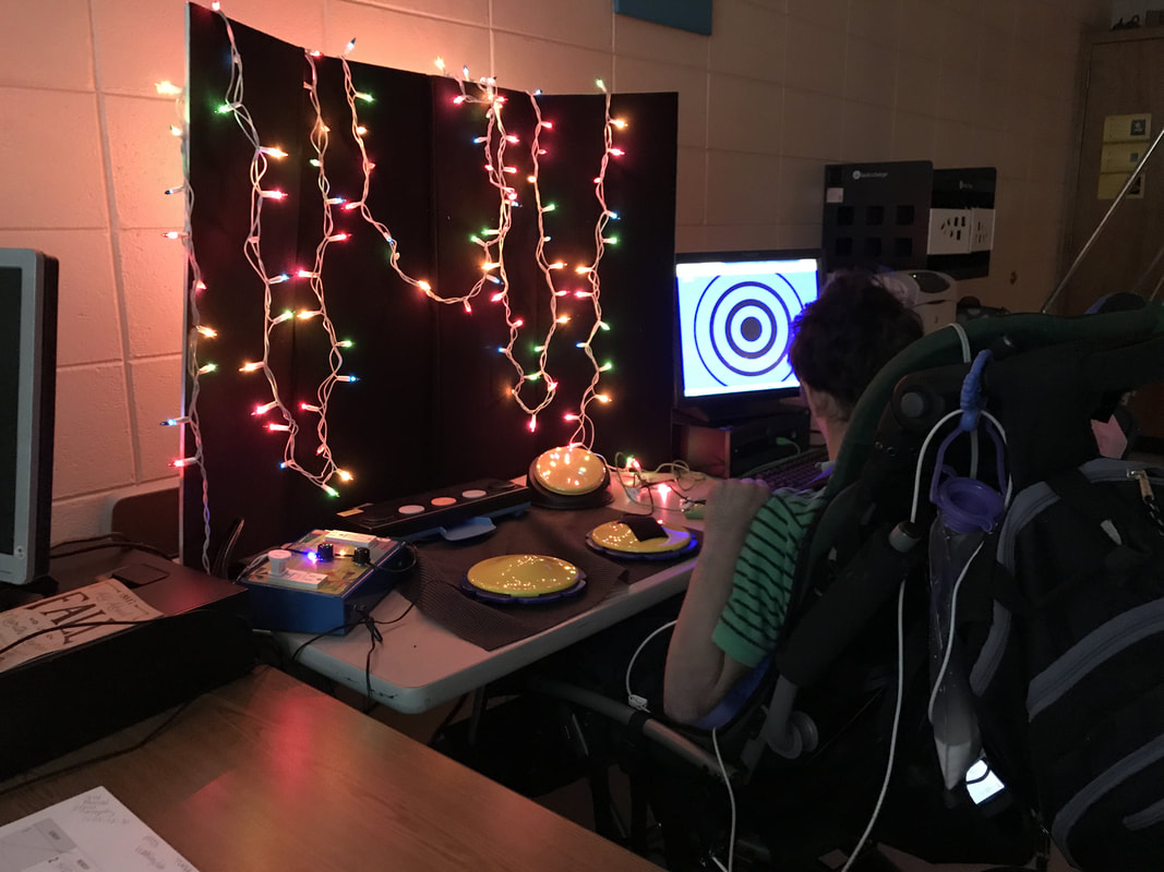 Picture of multicolored lights suspended on APH Invisiboard on table. APH Power Select and switches placed in front of student who is viewing a HelpKidzLearn game on computer.